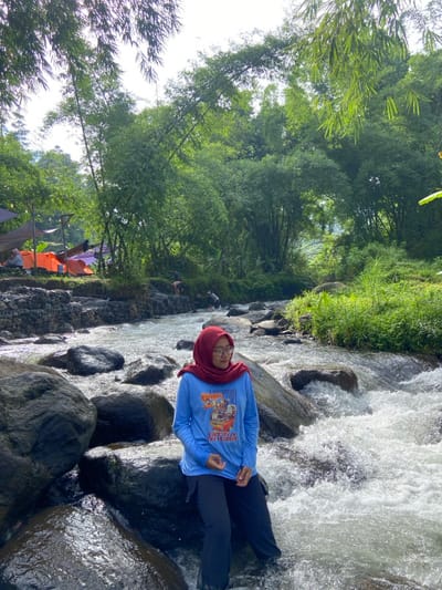 A woman in a hijab, blue long-sleeve shirt, and dark pants stands on rocks in a shallow, fast-moving stream surrounded by lush green vegetation. Tall bamboo trees are visible on the left side of the frame. Tents can be seen pitched near the stream, suggesting a campsite. The woman faces the viewer, partially immersed in the water. The surrounding environment is predominantly green with rocky elements. The lighting is bright and natural.