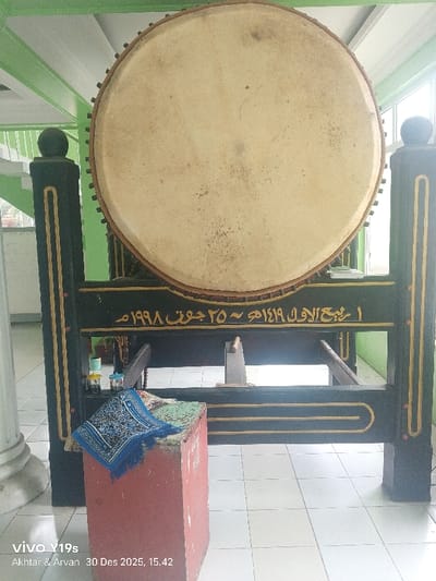 A large, circular bedug (drum) dominates the center of the image, positioned within a mosque or religious building in Jagapura Lor, Indonesia. The drum features a light, worn hide surface secured by numerous dark studs along its perimeter. It rests on a robust, dark-colored wooden stand adorned with gold-colored decorative trim. A horizontal beam on the stand displays gold Arabic script translating to "1 Rabi' al-Awwal 1419 H ~ 25 June 1998 AD."
In the foreground, to the left, a reddish-brown rectangular pedestal or box stands on a white tiled floor. A folded blue and white patterned prayer mat with fringed edges is placed on top of this pedestal. Behind the pedestal and beneath the drum, several small, colorful containers or bottles are visible on a lower ledge.
The background shows structural elements painted in a light green color. To the far left, a white column is partially visible. The floor throughout the visible area consists of clean, white square tiles. Natural light appears to enter from an unseen opening or window in the upper background. No individuals or active interactions are depicted.