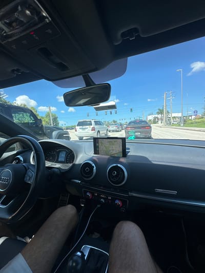 The image depicts a forensic view from the driver's seat of a dark-colored Audi vehicle, stationary at the intersection of Burnt Store Road and Veterans Parkway in Fort Myers, Florida, United States. The time is daytime, under a clear blue sky with scattered white cumulus clouds.
**Scene Elements:**
* **Driver:** The lower legs of an individual, likely male, wearing white shorts, are visible, indicating the driver's position. No other identifying features are visible.
* **Vehicle Interior (Audi):** The black dashboard and steering wheel, bearing the Audi logo, are prominent. The instrument cluster indicates a speed of "0". A mobile device or dedicated navigation unit is mounted on the dashboard, displaying a map of the immediate Fort Myers area, clearly showing "Burnt Store Rd" and "Veterans Pkwy". A charging cable extends from a device to the center console. A white rectangular object, consistent with a SunPass toll transponder, is attached to the windshield below the rearview mirror.
* **Traffic:** The Audi is queued in multiple lanes of traffic. Directly ahead is a black Dodge Challenger, with a small, light-blue object (possibly a toy or air freshener) visible on its lower rear bumper. To the right of the Challenger is a white Toyota Highlander SUV. A dark-colored Jeep Wrangler is positioned to the immediate left of the Audi. Other vehicles are present further down the road.
* **Roadway & Intersection:** The road is multi-lane asphalt with clear white lane markings. Traffic lights at the intersection are visible and appear green for through traffic.
* **Environment:** Tropical vegetation, including palm trees, lines the roadside. Utility poles and overhead power lines are visible on both sides of the road. Commercial buildings, including what appears to be a hotel structure, are visible in the background to the right. The overall impression is a typical sunny day in Southwest Florida.