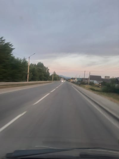 The image depicts a view from a vehicle traveling on a paved road, likely in or near Mayma, Russia, given the context. The road is asphalt with white lane markings. On the left side of the road, there is a guardrail and a dense line of trees. On the right side of the road, the terrain slopes slightly upwards, and there are several buildings that appear to be part of a rural settlement, with roofs of varying colors and materials. Power lines and utility poles are visible throughout the scene. The sky is overcast with soft, diffused light, suggesting either dawn, dusk, or a heavily clouded day. The image is slightly blurred due to motion.