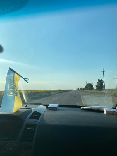 The image depicts a view from inside a vehicle looking out through the windshield. In the foreground, on the dashboard, is a small Ukrainian flag attached to the windshield with a suction cup. To the right of the flag are two packs of cigarettes. The road ahead stretches into the distance, lined by telephone poles and trees. On either side of the road are fields, with a large field of sunflowers visible to the left. The sky is clear and blue. The location is stated to be in the city of Maiske, Ukraine.
