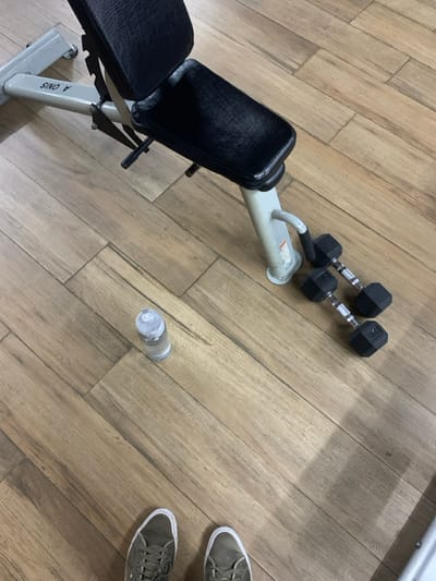 The image shows a weight bench and dumbbells in a gym located in Kecamatan Kartasura, Indonesia, at coordinates -7.567, 110.757. The bench is made of metal and has a black padded seat. It is in the upper left portion of the image. Three black dumbbells are lying on the floor next to the bench. One dumbbell is slightly closer to the bench than the other two. A water bottle is sitting on the floor next to the dumbbells. The floor is made of wood laminate with a brown color scheme. A pair of olive green sneakers are at the bottom center of the frame, partially visible. The room is brightly lit with the light source emanating from the left side of the frame. No people are present.