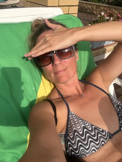 A woman is lying on a green towel on a lounge chair outdoors. She is wearing sunglasses and a patterned bikini. Her left hand is resting on her forehead, shielding her eyes from the sun. The background shows a stone wall and some plants, suggesting a residential or recreational area. The image is taken from a high angle, looking down at the woman. The location is Yaroslavskaya, Russia.