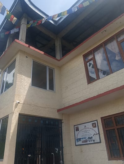 The image shows a two-story building with a textured, light-colored exterior. Prayer flags are strung across the top of the structure, which appears to be under construction or renovation, with exposed beams and corrugated metal roofing visible. The building has several windows, some with wooden frames and others with metal frames. There is a black metal gate on the ground floor. A sign on the wall reads "Buddhist Cultural Society of Key Gompa" and lists His Holiness the Dalai Lama as Patron and Ven. T.K. Lochen Tultu as President. There are two small images of individuals on the sign. A security camera is mounted on the exterior wall near the upper corner of the ground floor. The scene is outdoors, under a bright sky.