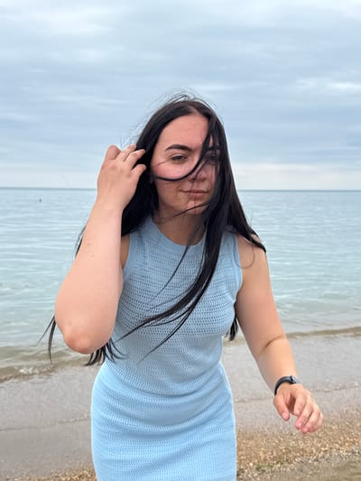 A young woman with long, dark hair is captured in a selfie by the sea. She is wearing a light blue, sleeveless, knit dress. Her left arm is extended, and she is touching her hair with her right hand, which is resting on her head. She is wearing a black band on her left wrist, possibly a fitness tracker. The background is a serene body of water under a cloudy sky, suggesting a cool or overcast day. The shore to the right appears sandy and pebbly. The image is oriented vertically, with the woman leaning into the frame. The lighting is soft, indicating it might be early morning or late afternoon. The overall mood is relaxed and contemplative. There is no visible text in the image. The location is identified as Derbent, Russia.