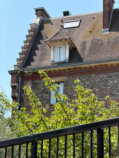 This is a vertical, eye-level, medium shot of a stone and brick building with a dark tiled roof in Courbevoie, France. The building has a steep roof with multiple dormer windows, chimneys, and decorative architectural elements. The stonework on the lower portion of the building is rough and irregular, while the upper sections feature brickwork and a decorative frieze. Green foliage from trees partially obscures the lower part of the building. A black metal railing is in the foreground, creating a barrier. The sky is clear and blue. The image captures a sunny day with strong shadows cast by the architectural features and foliage.