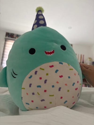 A close-up shot of a light turquoise plush toy in the shape of a shark. The shark has a white belly patterned with colorful confetti shapes. It has a small, spotted party hat on its head and a toothy grin. The background is blurred but suggests a residential setting. The image does not contain any information about a location in Casa Blanca, Mexico.