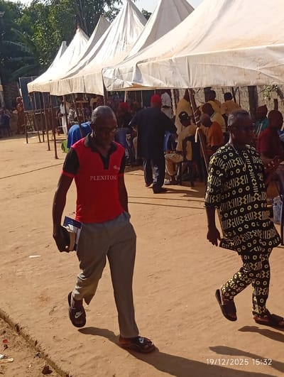 An outdoor scene shows several tents providing shade for seated people. Two men in the foreground are walking. The first man wears a red shirt, grey pants, and sandals, carrying what appears to be books. The second man is dressed in a patterned black and yellow outfit. The ground is a dirt road with a timestamp "19/12/2025 15:32" visible in the corner. Trees and foliage are visible in the background.