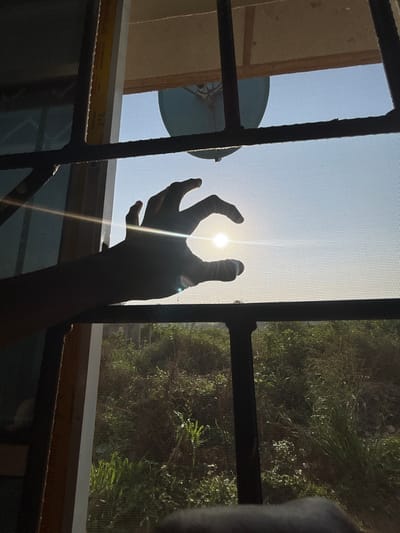 The image depicts an outdoor scene viewed from inside a structure in Debasco, Nigeria, through a window. The window is equipped with dark-colored, vertical and horizontal metal security bars and an insect screen. A human hand, silhouetted against the light, occupies the central foreground, positioned with fingers slightly curved as if cupping or reaching towards the sun. The sun is a bright, circular light source visible through the hand, low in the sky, producing a prominent lens flare across the image. Outside the window, above the horizon, a light blue satellite dish is visible against a clear, light blue sky. Below the horizon, a dense, uncultivated area of green and brown vegetation, including tall grasses and shrubs, is present. A vertical section of a light-colored window frame is partially visible on the far left of the image.