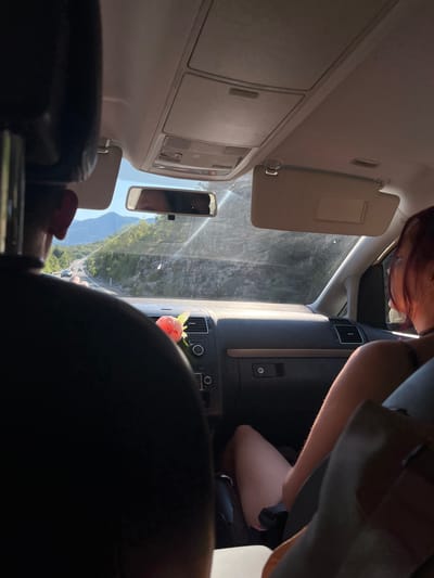 The image depicts the interior of a car, likely in or near Gornji Ulići, Montenegro, with a view of the road and surrounding landscape through the windshield. Sunlight streams into the car, creating glare on the windshield and dashboard.
In the foreground, the back of a person's head and shoulder are visible on the left. On the right, a person with reddish hair is seen from the side, with their legs resting in the passenger seat. A flower, possibly a peony, is placed on the dashboard.
The dashboard features an air vent, a car stereo system, and a glove compartment. The rearview mirror reflects the driver's face. Outside the car, a winding road is visible with a few cars traveling on it. The landscape is mountainous, covered in green vegetation, and the sky is clear blue. The overall impression is of a scenic drive on a sunny day.
