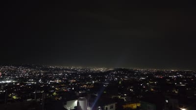 The image depicts a nighttime view of Tonalá, Mexico. The city sprawls across the landscape, with numerous lights creating a bright and dense pattern against the dark background. The sky is dark. In the foreground are the tops of buildings and structures.