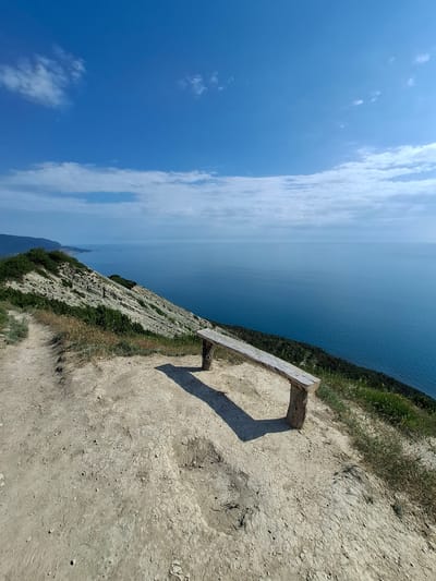 A wooden bench sits on a dirt path overlooking the sea in Varvarovka, Russia. The bench is positioned facing the ocean, with a vast expanse of blue water stretching to the horizon. The path is unpaved and dusty, with dry grass and sparse vegetation growing along the edges. The terrain slopes downward towards the sea, revealing layers of rock formations on the cliff face. In the distance, mountains can be seen along the coastline under a bright blue sky dotted with fluffy white clouds. The overall scene is one of natural beauty and tranquility.