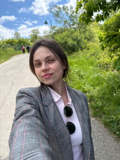 A woman is posing for a selfie in what appears to be a park in Sremska Kamenica, Serbia. She is wearing a gray plaid blazer over a light pink shirt and sunglasses are hooked on her shirt. Behind her is a paved path flanked by grass and trees. Further in the background, there are other people walking on the path under a blue sky with clouds. A lamp post can also be seen in the background.