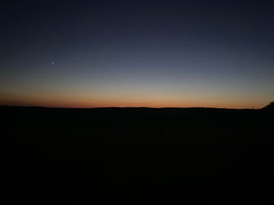 This media file captures a twilight scene in Negrești, Moldova. The image depicts a vast, dark landscape under a gradient sky that transitions from deep navy at the top to a soft orange and yellow near the horizon. A single, bright star or planet is visible in the upper left quadrant of the frame. The horizon line is silhouetted against the lighter sky, suggesting a hilly or uneven terrain. The foreground is almost entirely black, obscuring most details of the ground, but a faint hint of vegetation or a fence post can be discerned in the darkness. The overall atmosphere is serene and tranquil, indicative of dusk or pre-dawn. No people, text, or specific activities are visible. The setting appears to be rural, with the vastness of the sky and land dominating the composition.