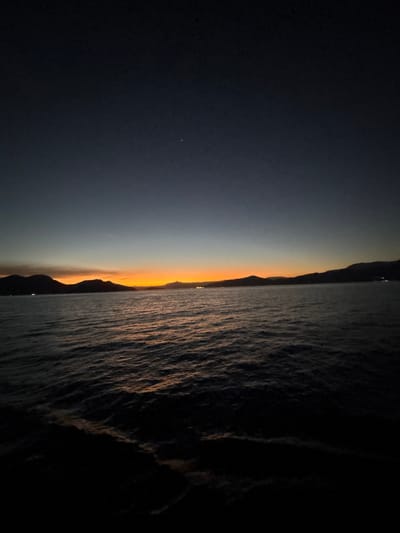 The image depicts a tranquil scene of the sea at dusk or dawn, with a clear horizon and a gradient of colors in the sky. The sky transitions from a deep dark blue at the top to lighter shades of gray and then a vibrant orange and yellow band near the horizon. Silhouetted mountains line the distant shore, some with faint lights suggesting human presence. The water in the foreground is dark and choppy, reflecting the dim light of the sky. A single bright star is visible in the upper right portion of the sky. The location is identified as Angra dos Reis, Brazil, suggesting a coastal environment. There are no people or overt actions depicted; the scene is one of natural beauty and stillness.
