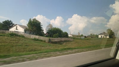 The image shows a rural scene in Krasnoshchekovo, Russia, viewed from inside a vehicle. A weathered wooden fence runs along the foreground, separating a grassy slope from a cluster of buildings and trees in the middle ground. On the left, a white house with a red roof and a small dormer window is partially visible behind the fence. To the right of this house, a dense line of trees and bushes obscures the view of further structures. Further to the right, another white building with a dark roof stands amidst greenery, with additional buildings and what appears to be a light-colored utility pole visible in the distance. The sky above is a pale blue, dotted with fluffy white clouds. The road in the foreground is asphalt and shows motion blur, indicating the image was taken while the vehicle was moving. Reflections from the vehicle's window are visible on the right side of the image.
