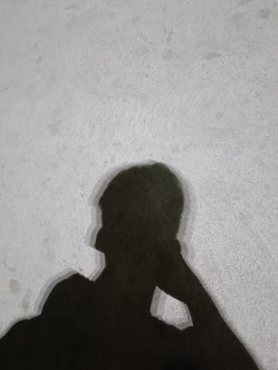 The image shows the shadow of a person leaning their head on their hand, cast on a textured, light-colored surface, possibly concrete or stucco. The shadow is dark and has a soft outline, indicating a diffused light source or distance. The surface appears to be outdoors and somewhat gritty, with subtle variations in tone and small specks. The composition is minimalist, focusing on the silhouette of the person and the texture of the surface. The scene is devoid of any identifiable landmarks or urban elements that would specifically place it within Almalyk, Uzbekistan.