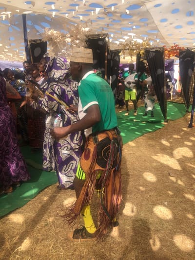 The image captures an outdoor cultural gathering in Kerang, Nigeria, under a white canopy with decorative cut-outs, allowing dappled sunlight to filter through. The ground features patches of green carpet and dry, light-colored earth.
In the foreground, a man wearing a light green t-shirt with the partial text "ALL PROGRESIST PA" (likely "ALL PROGRESSIVES PARTY") and a woven, light-colored hat is prominent. He is leaning forward, actively engaged in playing a drum-like instrument with a stick. He wears elaborate traditional attire around his waist and lower back, characterized by brown leather, fringes, and decorative patterns. Directly behind him, a woman in a purple and white patterned dress is visible.
Several other individuals are present in the mid-ground and background, some seated and others standing. Their attire varies, including traditional African patterns and casual wear. Black fabric partitions or decorative elements are visible further back. The overall scene suggests a vibrant event, possibly a musical performance or celebration, taking place during the day under bright, sunny conditions.