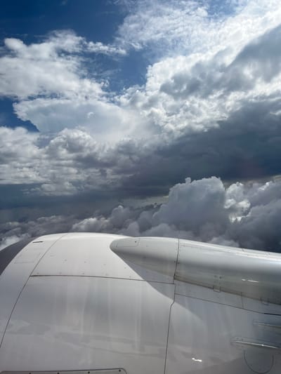 The image shows a view from inside an airplane, looking out the window. The foreground is dominated by the white engine casing of the aircraft wing. Beyond the wing, the scene depicts a sky filled with various cloud formations, ranging from fluffy white clouds to darker, more ominous-looking storm clouds. A patch of blue sky is visible in the lower left corner. There is no indication of the city of Rustavi, Georgia, nor are there any people, subjects, objects, actions, or interactions visible other than the plane and the clouds.