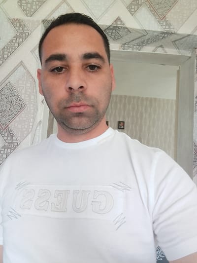 The image shows a man wearing a white t-shirt with the "Guess" logo. He is standing indoors, facing the camera. The background features patterned wallpaper. A reflection in a doorway to the right shows a different patterned wallpaper and what appears to be a framed picture on the wall. The overall scene suggests a domestic indoor environment. The location is identified as Bukovlak, Bulgaria.