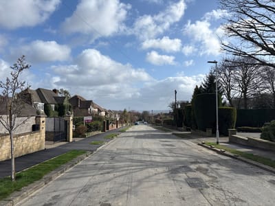 This forensic description details a static daytime scene on a residential street in Kirklees, United Kingdom, likely within the Honley/Holmfirth area, under a partly cloudy sky with discernible sunlight and shadows.
The asphalt road, presenting surface wear, cracks, and multiple rectangular repair patches, extends centrally into the distance with a gentle downward slope. On the left side of the street, a continuous pavement is bordered by a narrow grass verge and a stone wall. Behind this wall, a row of traditional-style houses, featuring stone facades and dark, pitched roofs, is visible. The closest house on the left displays a prominent stone wall, a dark metal gate, and stone pillars. Further down on the left, a purple "For Sale" sign from "Roger J. Speight" is positioned within a garden area.
The right side of the street also features a pavement and grass verge, with properties largely obscured by tall, well-maintained dark green hedges. A modern, tall, grey street light with a rectangular LED fixture stands on the right pavement in the mid-ground. Beyond the hedges and street light, bare deciduous trees are visible against the sky. Overhead power lines or telephone lines traverse the upper portion of the frame. Two light-colored vehicles are visible in the distance on the left side of the road. No individuals are present in the scene. The distant horizon suggests open terrain or hills.