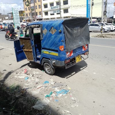 A blue, three-wheeled auto-rickshaw (tuk-tuk) with its passenger doors open is parked on the shoulder of a paved road in Ongata Rongai ward, Kenya. The vehicle, appearing worn with faded paint and a patched black canvas roof, displays license plate KWB 771R on its rear. To its immediate left, a patch of dirt is heavily littered with various pieces of trash, including plastic bags and debris.

The road features a painted white median line and a broken yellow line indicating traffic flow. In the background, a person on a red motorcycle, wearing a dark helmet and backpack, drives left to right. Further back, multiple parked cars, predominantly white, are visible on the far side of the road.

Several multi-story commercial buildings line the street. On the left, a building prominently features signs for "OUTFIT SUNSHINE THE WHOLISTIC LABORATORY" offering "X-RAY ULTRASOUND CT-SCAN MRI DENTAL OPTICAL." A "NOW OPEN" banner is also visible. On the right, a building displays a "MARITE STORES" sign. The sky is partly cloudy, and the lighting suggests daytime conditions.