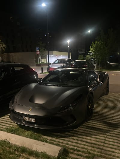 The image captures a nighttime scene in what appears to be a parking area in Dübendorf, Switzerland. The primary subject is a dark, matte-finished sports car, likely a Ferrari, parked on a permeable pavement surface featuring concrete strips interspersed with grass. The car's sleek design and dark wheels with yellow accents (possibly brake calipers or center caps) are visible.
The time of day is clearly night, with multiple streetlights illuminating the scene, casting reflections and shadows. The background shows other parked vehicles and distant buildings, barely visible through the darkness, along with some trees.
A prominent detail is the Swiss license plate on the rear of the main car, which reads "ZG-98036". "ZG" corresponds to the Canton of Zug, providing a specific geographical cue within Switzerland, aligning with the provided location context. In the mid-ground, a blue road sign with a white "P" symbol for parking is visible, next to a red circular sign with "18t" inside, likely indicating an 18-ton weight limit. Another small, red circular sign is partially obscured.
No people are visible, and no active event is taking place; the cars are stationary. The overall atmosphere is quiet, typical of a parked area at night.