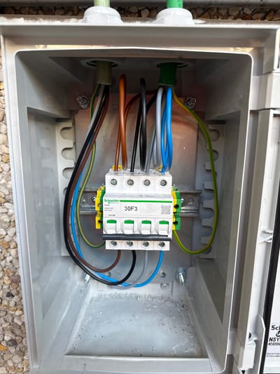 The image shows an open, gray electrical junction box with a white plastic exterior, mounted outdoors. The box contains a DIN rail with a Schneider Electric 30F3 circuit breaker. Several electrical wires, in colors including brown, black, gray, blue, and yellow-green, enter the box through two green cable glands at the top and connect to the circuit breaker. The interior of the box appears dusty. The box is positioned outdoors, with a gravel-like ground visible in the background and to the left. A white label is attached to the right side of the box, partially obscured, with visible text including "Sch" and "NSYC". The setting is identified as Zwijndrecht, Belgium.