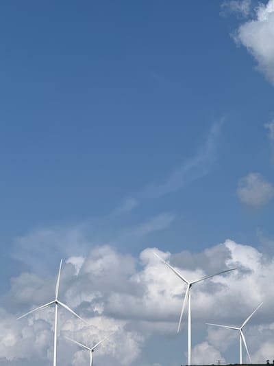This image depicts a wind farm against a blue sky with scattered clouds. Several white wind turbines are visible, with their blades extended. The turbines are positioned on a slight incline, suggesting a hilly terrain. In the background, a faint power line structure can be observed. The scene is tranquil and evokes a sense of natural energy generation. The image is oriented vertically.