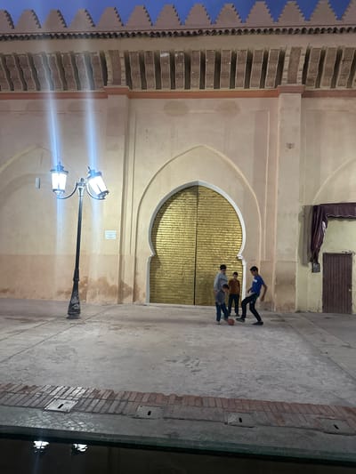 The image captures an exterior shot within the Mechouar Kasbah, Morocco, at night. Four boys are seen playing with a ball in front of a large, ornate, arch-shaped golden door. The architecture features traditional Moroccan design, with intricate patterns and textures visible on the building's facade. A vintage-style lamppost illuminates the scene, casting light on the stone pavement. The overall atmosphere is one of everyday life unfolding within a historical setting.