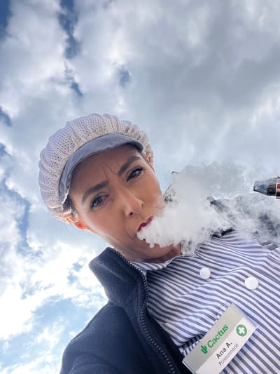 An extreme low-angle outdoor portrait shows a woman exhaling a prominent cloud of white vapor from her mouth against a bright sky with scattered clouds. The woman, identified by a name badge as "Ana A." from "Poissonnerie" (fishmonger) at "Cactus," is wearing a white perforated mesh cap, a black zippered fleece jacket over a blue and white striped shirt. Two round white objects, possibly buttons, are visible on her shirt. Her lips are covered in red lipstick, and her eyes, with visible makeup, are directed towards the viewer. The "Cactus" name tag, featuring a green cactus logo and a white cross icon on a green background, indicates her affiliation with the Cactus supermarket chain. While the image does not show specific city landmarks, the identification with "Cactus", a major supermarket in Luxembourg, and the existence of a Cactus store in Roodt-sur-Syre, Luxembourg, implies the scene is associated with or near that location.