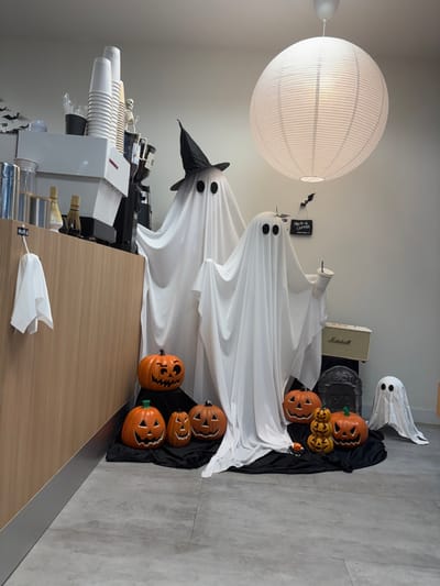 This image depicts a Halloween-themed interior scene within a commercial establishment in Tambon Hua Toei, Thailand. The primary focus is a display of three white fabric ghost figures positioned on a black draped cloth on a light gray tiled floor. The largest ghost, wearing a black pointed witch's hat, stands center-left, with a medium-sized ghost to its right holding a white disposable cup with a straw. A smaller, plain ghost is situated further right. Surrounding these figures are approximately seven orange plastic or ceramic jack-o'-lanterns of varying sizes, some carved with faces. A stack of three smaller, uncarved pumpkins is also present on the black cloth, along with a small black spider decoration and a gray tombstone prop.

In the background, to the left, a light-colored wooden counter features stacks of white disposable cups, a black container, a small skeleton figurine, and commercial coffee-making equipment, including components of an espresso machine and two small glass bottles with whisks. A small white fabric ghost shape hangs from the front of this counter. The walls are a plain light gray. A large, ribbed white spherical paper lantern hangs from the white ceiling. Black bat cutouts adorn the wall above the ghost figures, along with a small black sign partially obscured but appearing to read "MUM-A COFFEE." To the far right, behind the smaller ghost and tombstone, a cream-colored Marshall amplifier is visible. The scene is brightly illuminated, indicating an indoor setting. No human figures are present.