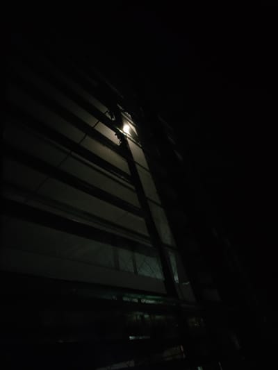 This is a nighttime photograph taken from a low angle, looking up at a building facade with horizontal lines and a tall, dark vertical structure in front of it. The scene is very dark, with a single bright light source visible, casting a soft glow and shadows. The building appears to be constructed with large panels, and there are horizontal elements that suggest balconies or structural beams. The vertical structure in the foreground is partially obscuring the building and has a mesh-like pattern visible through some of its openings.
The time of day is clearly night, and the atmosphere is dark and somewhat mysterious due to the limited illumination. There are no people or animals visible. The overall impression is one of an urban setting, possibly industrial or residential, with a focus on architectural lines and shadows. The light source seems to be an artificial light, possibly a lamp or a security light. The texture of the building's surface is grainy, indicating the image was taken in low light conditions, potentially with a smartphone. There is no visible text in the image.