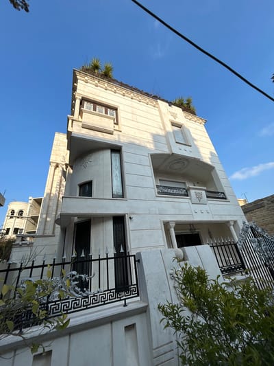 The media file showcases a multi-story, light-colored building, likely a residential property, under a clear blue sky. The building's exterior is adorned with ornate architectural details, including light-toned stone or marble cladding, decorative moldings, and carved elements.
Prominent features of the facade include a large, central arched recess on an upper floor with a circular relief carving, flanked by classical columns and patterned panels. Balconies and windows are secured with elaborate black metal railings, featuring intricate scrollwork, Greek key patterns, and pointed finials. Green plants, possibly palm varieties, are visible on the rooftop or upper terraces, providing a natural accent. A leafy green bush or tree also partially frames the building on the right side.
The sky is a bright, unblemished blue, indicating clear and sunny weather. Sunlight illuminates the building, creating subtle shadows consistent with a high sun angle during morning or afternoon. A black cable or power line diagonally traverses the upper left of the frame. In the background of some views, another building with arched windows and a dome-like structure, possibly under construction, can be seen. A small security camera is discreetly placed on a wall.
The image is captured with a tilted perspective, rotating the building counter-clockwise. No people or activities are depicted, focusing solely on the architectural subject. No visible text is present in the scene. The location context for this building is Aleppo, Syria.