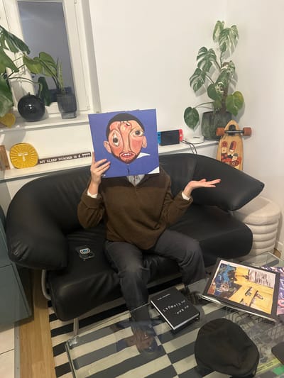 A person sits on a black leather chair, holding a vinyl record with a cartoonish portrait over their face. The person is wearing a brown sweater and gray pants. Next to them is a glass table with books and a black baseball cap. Behind the chair is a skateboard, a Nintendo Switch, and a monstera plant. In the bottom left corner of the image, there is a window with another monstera plant in front of it.