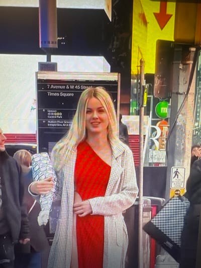 The image shows a woman with blonde hair, wearing a red one-shoulder dress and a patterned jacket, standing on a street. She is smiling and looking to her right. To her left, another person in dark clothing is visible. Behind the woman, a sign reads "7 Avenue & W 45 Street Times Square". Various signs and structures suggesting an urban environment, possibly Times Square in New York City, are visible in the background, including an M&M's store sign. There is no indication of Chorzów, Poland. The lighting suggests it is daytime.