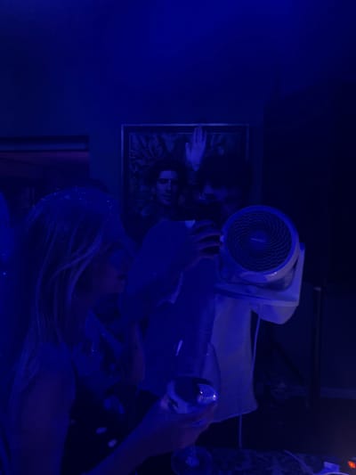 In a dimly lit room in Pozuelo de Alarcón, Spain, bathed in blue light, a woman with blonde hair is seen in profile, looking down. She is holding a glass. In front of her, a person is holding a white portable fan, with their face partially obscured. Behind them, a framed artwork hangs on the wall, depicting a person's face and a hand. Another person's face is visible in the background, looking towards the camera. The environment suggests a social gathering or party.