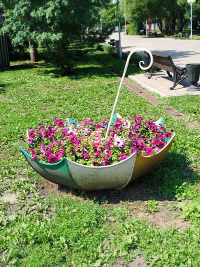 An outdoor scene in Yalutorovsk, Russia, features a flowerbed shaped like an umbrella, filled with vibrant purple and pink petunias. The umbrella-shaped planter is a weathered, hollowed-out object with a white and yellow exterior and a green interior, resting on a grassy area. A white, curved metal pole, reminiscent of an umbrella handle, extends from the planter, curving upwards and to the right. In the background, a paved pathway runs through a park lined with trees. A dark-colored park bench with a wooden seat and metal legs is situated on the pathway, accompanied by a black trash receptacle. Another similar trash receptacle is visible further down the path. The environment is characterized by green grass, mature trees casting shadows, and sunlight illuminating the scene.