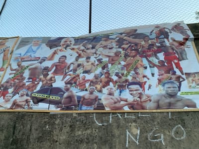 A large billboard is mounted on a concrete wall with a chain link fence in the background. The billboard features a collage of images of numerous men, mostly shirtless. Some of the men are wearing boxing gloves. The men's faces are generally serious or intense. There is a small car visible on the bottom left of the billboard. The car's license plate says "SOKOTO". The word "SOKOTO" is also printed on the billboard in large letters. The billboard is covered with text and slogans in Hausa and English. The wall is painted a beige color. Some graffiti is visible on the lower portion of the wall, including the word "NGO" scrawled in large, white letters. This scene is located in Sokoto, Nigeria, at coordinates 13.049, 5.238.