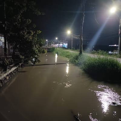At night in Brangsong, Indonesia, a narrow, dark canal reflects the light from street lamps lining a road. The canal banks are lined with vegetation. A building is seen on the left of the image and stores are on the right. There appears to be a bridge over the canal in the distance, people are on the bridge.
