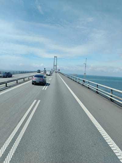 The image depicts a view from a vehicle driving across a large suspension bridge on a clear day. The bridge spans over a calm body of water, likely the sea, with a hazy horizon and distant wind turbines visible. The sky is predominantly blue with scattered white clouds.
Several vehicles are present on the bridge, including a dark gray Mercedes-Benz sedan in the foreground, a black sedan to the left, and several vans and trucks further ahead. The road is asphalt with clear white lane markings. Metal railings line the sides of the bridge, separating the roadway from the water. A tall pole with electronic equipment is visible on the right side of the bridge, possibly a weather station or traffic sensor. The overall scene suggests a normal day of traffic crossing the bridge.
The lighting indicates it is daytime, likely midday given the brightness of the sky. The weather appears pleasant and sunny. The presence of the suspension bridge and the vast expanse of water strongly suggests a coastal or strait crossing. The distant wind turbines are a key indicator of the location being Denmark, specifically near Korsør where such structures are common. No discernible text is visible in the image, apart from license plates on vehicles which are too blurry to read.