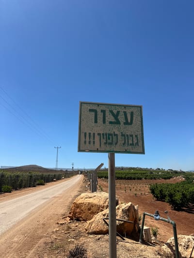 The image displays a sunlit outdoor scene in the Galil Elyon Regional Council, Israel, featuring a dirt road and a prominent warning sign. The sky is a bright, clear blue, indicating a sunny day, likely mid-morning or early afternoon.
A weathered, light green rectangular sign stands on a metal pole to the left of the dirt road. The Hebrew text on the sign is faded but legible. It reads:
"עצור" (Atzor) – meaning "STOP"
"גבול לפניך!!!" (Gvul Lefanecha!!!) – meaning "Border Ahead!!!"
The dirt road itself extends into the distance, flanked by agricultural land. On the left side, rows of cultivated green trees, possibly an orchard or vineyard, are visible, bordered by a fence. On the right, large, light-colored rocks are interspersed with dry vegetation, and a section of an irrigation pipe system with a valve is attached to the rocks. Further along both sides of the road, more fields with green foliage stretch towards the horizon, where low hills are visible in the background. Power lines are strung across the landscape, connecting distant utility poles. The scene is devoid of people, conveying a sense of quiet rurality near what is indicated as a border or frontier.