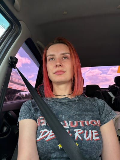 A fair-skinned woman with red hair is wearing a gray t-shirt with distressed lettering and yellow stars. She is wearing a seatbelt in the passenger seat of a car. Outside the window, there is a view of a building under a sky with pink-tinged clouds. The location is Горняк, Russia.