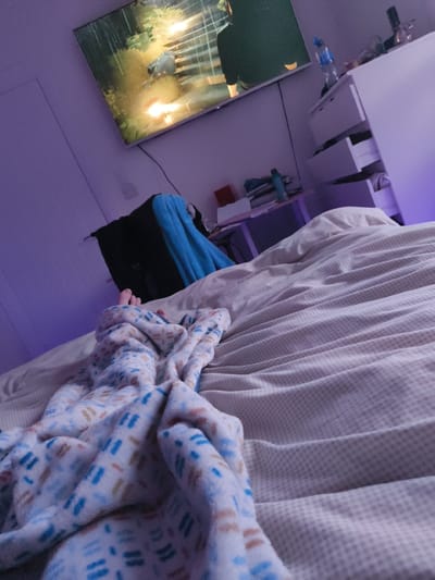The image shows a person's feet and legs wrapped in a patterned blanket. They are lying on a bed with white and beige checkered bedding. There is a flat-screen television on the wall, playing a scene from what appears to be a game or movie. A black and blue bag lies on the bed near the person's feet, and a small desk is visible in the background. The person is looking directly at the camera. The room is a bedroom in a private residence. The location is Newcastle, United Kingdom, at the coordinates 54.207 latitude and -5.9 longitude. The scene is set in a quiet bedroom, likely at night or in the evening. The person appears to be resting or relaxing in the bedroom. The lighting is soft and warm, with a faint purple glow emanating from under the bed.