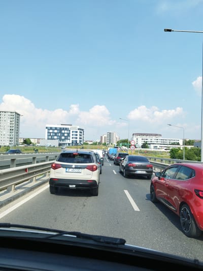 The image depicts a traffic jam on a multi-lane road, likely a highway or elevated road, with several cars visible. The scene is set in Bragadiru, Romania, indicated by the building in the background with "DIMRI" and a Romanian phone number visible. The sky is bright blue with scattered white clouds. On the left side of the road, a tall, modern apartment building is present, and further in the distance, other buildings and greenery can be seen. On the right side of the road, there are traffic signs, including a speed limit sign indicating 50. The cars are bumper-to-bumper, suggesting a significant delay. The perspective is from inside a vehicle, looking forward through the windshield, with the dashboard and windshield wipers partially visible. The vehicles are a mix of makes and models, with a beige Suzuki Vitara in the foreground on the left, a red Renault Clio on the right, and various other sedans and SUVs in the queue ahead. The road surface is asphalt, with white lane markings. The overall impression is a typical urban traffic congestion scenario on a clear day.