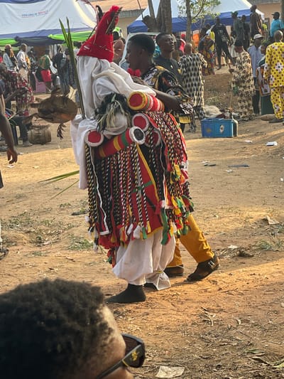 An outdoor scene in Umueze, Nigeria, depicts a public gathering with a central focus on a masquerader. The masquerader, a person wearing an elaborate traditional costume, is positioned slightly right of center, facing partially right. The costume features a conical red headpiece with red and green feather-like adornments, a white cloth covering the upper body, and a voluminous skirt-like lower body segment constructed from multiple strands of red, black, green, and yellow fabric strips with multicolored tassels. Several padded, cylindrical red, white, and black elements are attached to the upper part of the skirt. The masquerader holds a long green frond in their left hand and a dark, possibly wooden, skull-like or gourd object attached to a stick, which hangs from their left side. They wear white baggy trousers, dark socks on the left foot, and part of a mustard-yellow trouser leg with a dark, buckled shoe on the right foot.
Immediately to the right of the masquerader stands a young male attendant with short dark hair, wearing a patterned dark shirt and dark trousers. He appears to be observing or assisting the masquerader.
In the mid-ground and background, numerous individuals are gathered, dressed in various traditional patterned garments (including Ankara prints, camouflage-like patterns, and a yellow and white checkered outfit) and casual wear. They are standing on reddish-brown dirt ground.
Environmental elements include:
* Several white tents with blue trim are visible in the background, one bearing the text "UBASINACHI RENTALS."
* Trees and dense foliage are present in the distant background.
* A blue plastic crate is on the ground in the mid-right.
* A wooden drum is partially visible on the ground to the left of the masquerader.
* The ground is dry, dusty, and strewn with scattered debris and dry vegetation.
* The lighting is natural daylight, casting subtle shadows.
In the immediate foreground, out of focus at the bottom left, the top of a person's head with dark, curly hair and the upper frame of dark sunglasses are visible, indicating the perspective from within the crowd.