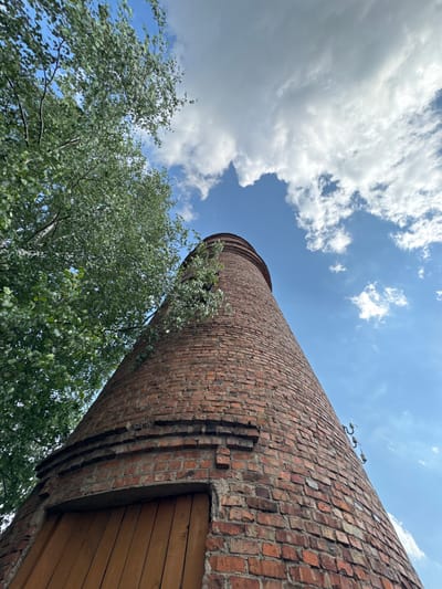 This is a low-angle, upward-facing shot of a tall, cylindrical brick structure, likely a chimney or a water tower, in Červėń, Belarus. The structure is made of red brick, showing signs of age and wear, with some bricks missing or broken. Green foliage from trees is visible in the lower left and right of the frame, partially obscuring the base of the brick structure.
The sky above is a vibrant blue, filled with large, fluffy white clouds. The sunlight suggests it is daytime, likely late morning or early afternoon, given the brightness of the clouds and the clear sky. There are a couple of metallic fixtures on the upper part of the brick structure, possibly electrical insulators or lighting fixtures. A section of the structure towards the right features wooden planks, indicating a doorway or an added enclosure.
The perspective emphasizes the height and scale of the brick structure, while the surrounding trees and the sky provide a natural setting. The weather appears to be pleasant and sunny, with a gentle breeze suggested by the way the leaves are slightly blurred. No people are visible in the image, and there is no visible text. The overall mood is one of rustic industrial architecture set against a beautiful natural backdrop.