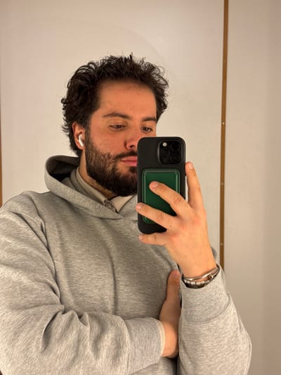 A male individual is depicted taking a mirror selfie in Gondomar, Portugal. He has dark, curly hair and a full beard, with a white earbud visible in his right ear. He is wearing a light grey hooded sweatshirt over a lighter-colored collared shirt. On his left wrist, he wears multiple bracelets, including a silver-toned cuff and a beaded bracelet. His right hand holds a black smartphone, equipped with a triple-lens camera system and an attached dark green wallet accessory, positioned to capture the reflection. His left arm is bent, with his hand resting against his torso, partially obscured by the sleeve of his sweatshirt. The background consists of a plain, light-colored wall with a vertical wooden strip or trim, featuring visible fasteners, located on the right side of the frame.