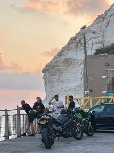 The image depicts a group of four young men and two motorcycles in the city of Mate Asher Regional Council, Israel. The scene is set at dusk, with a pastel-colored sky and a large white cliff face in the background. A body of water, likely the Mediterranean Sea, is visible to the left, bordered by a metal railing.
The men are dressed in casual attire. Two are standing near the railing, one leaning on it and holding a phone, while the other stands beside him. Two other men are standing closer to the motorcycles. One is holding a drink and looking towards the cliff, while the other is looking to the left.
Two motorcycles are parked in the foreground. The one on the left is black with blue accents and a yellow license plate. The motorcycle on the right is bright green with black accents and the word "Ninja" visible on its side. A black car is parked behind the motorcycles.
A brick building is situated against the base of the cliff, with a barred entrance. A yellow metal structure with a railing runs horizontally across the mid-ground. Signs with Hebrew and English text are visible on the building and near the yellow structure, one of which reads "NO ENTRY." The overall atmosphere suggests a casual gathering of friends in a scenic location.