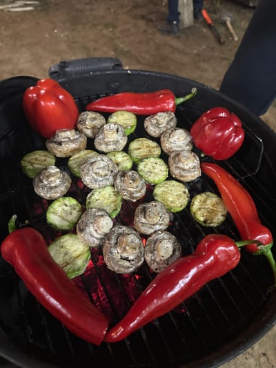 The image shows grilled vegetables on a round, black barbecue grill. The vegetables include whole red bell peppers and long, thin red chili peppers, as well as sliced zucchini and whole mushrooms. The grill is positioned outdoors, with a patch of grass or ground visible in the upper left corner. A person's leg and part of their upper body are visible in the upper left corner as well. The grilled vegetables are centered on the grill surface. The specific location is слобода Старая, Russia.
